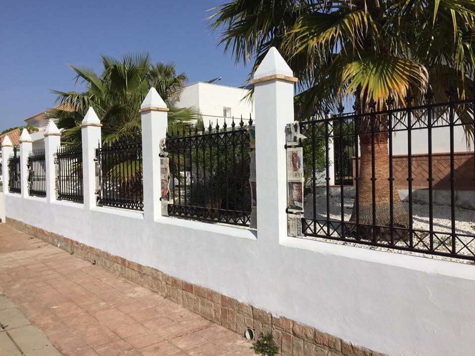 Valla perimetral blanca con verja de forja negra y palmeras